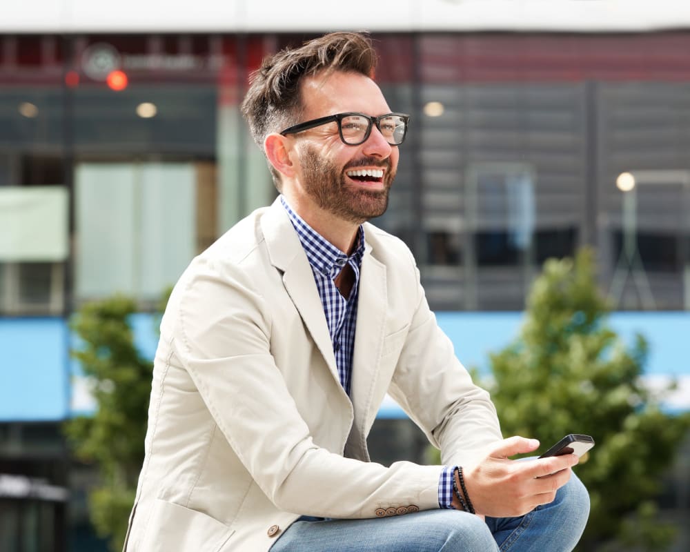 A white adult man is sitting outside and laughing. He's wearing glasses and holding a cell phone.