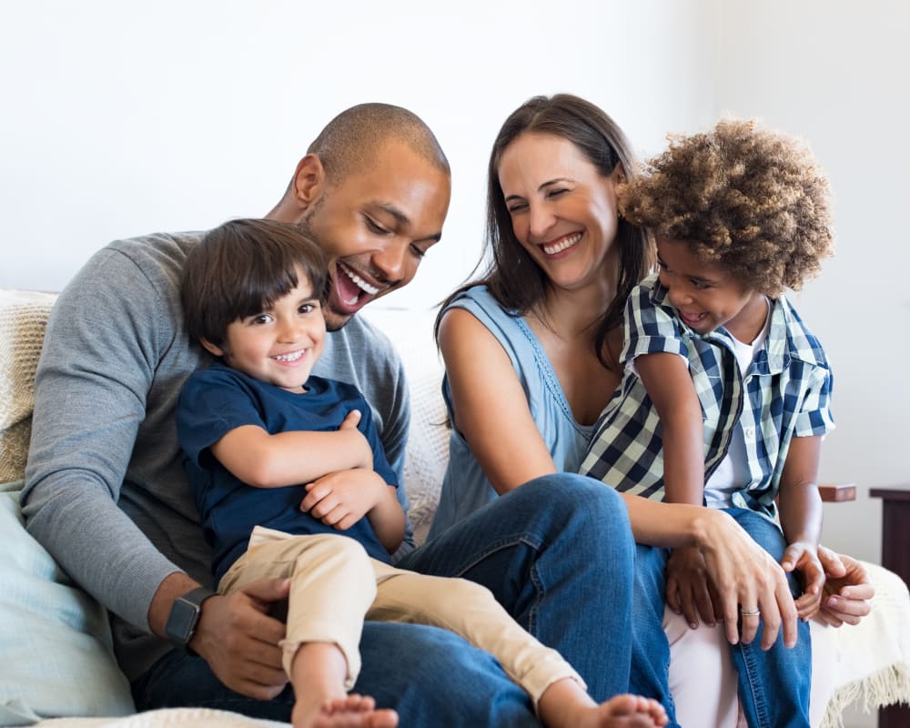 A family of four are smiling and snuggling together on the couch. Each parent has a young kid on their lap.