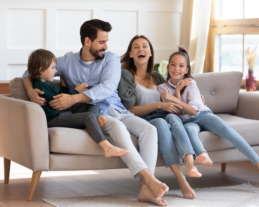 A young family of four is sitting on a couch smiling and laughing.