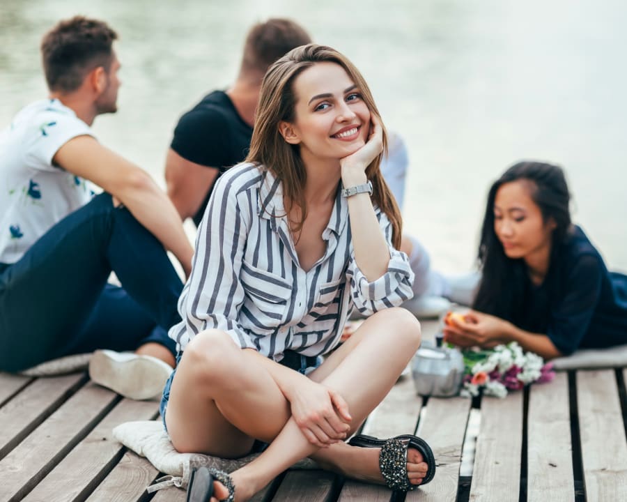 A young adult woman is sitting cross-legged on a dock, smiling with her head in her hand.