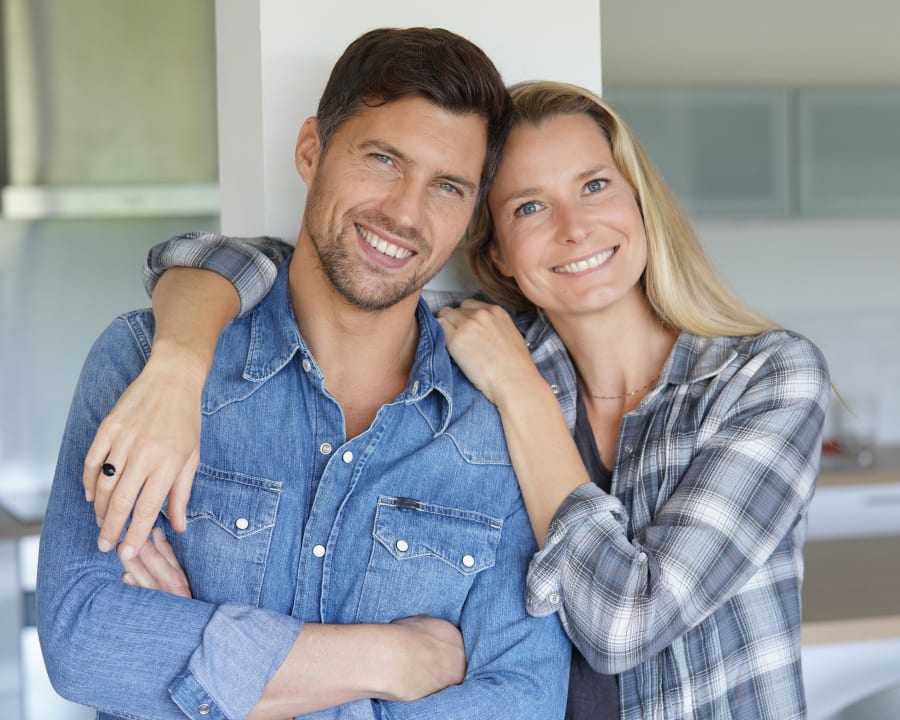 A man and woman are smiling, he has his arms crossed and she has one arm slung around his shoulders. Her other arm is placed on his shoulder closest to her.
