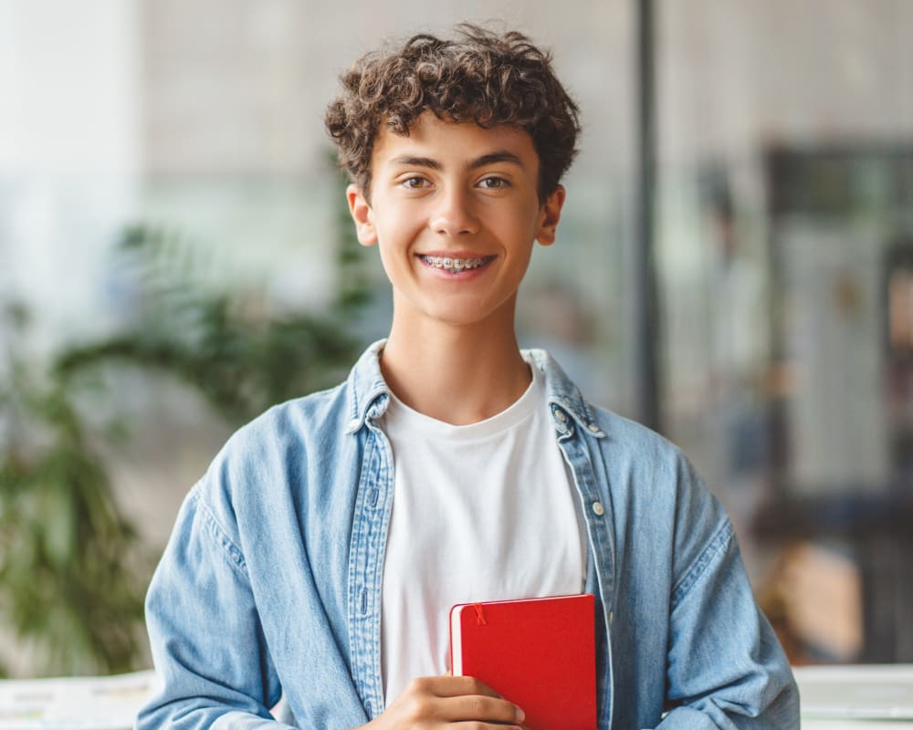 A teenage boy with braces is wearing a denim shirt and is holding a red book. He is smiling at the camera.